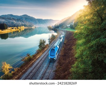 Aerial view of blue modern high speed train moving near river in alpine mountains at sunrise in summer. Top view of train, railroad, lake, reflection, trees in spring. Railway station in Slovenia