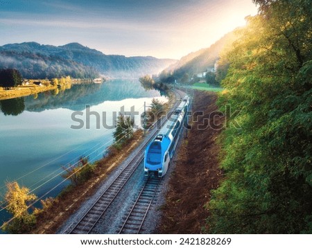 Aerial view of blue modern high speed train moving near river in alpine mountains at sunrise in summer. Top view of train, railroad, lake, reflection, trees in spring. Railway station in Slovenia