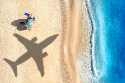 Aerial View Of Blue Sea, Airplane Shadow, Sandy Beach With Umbrellas At Sunset In Summer. Tropical Landscape With Blue Water, People, Shadow Of Flying Plane. Travel And Vacation In Islands. Top View