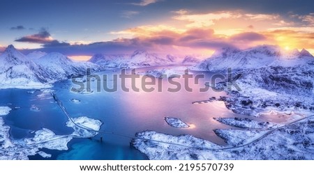 Aerial view of blue sea, snowy islands, mountains, sky with pink clouds at sunset in winter. Lofoten islands, Norway. Landscape with rocks in snow, reflection in water. Top view from drone. Nature