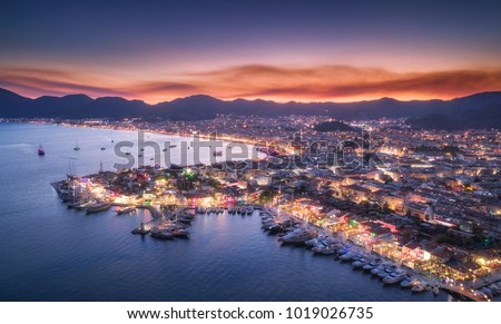 Aerial view of boats and beautiful city at night in Marmaris, Turkey. Landscape with boats in marina bay, sea, city lights, mountains, red sky, clouds at dusk. Top view from drone. Harbor at sunset