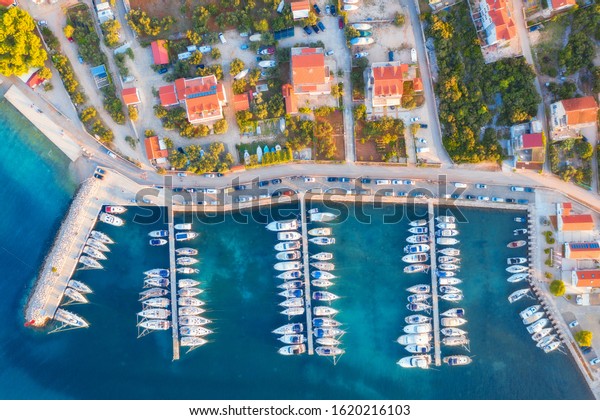 Aerial view of boats and yachts in port in old city\
at sunset. Summer landscape with houses with orange roofs,\
motorboats in harbor, clear blue sea, cars on the road. Beautiful\
architecture. Top view
