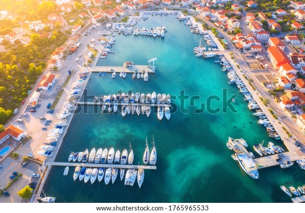 Aerial view of boats and yachts in port in beautiful\
old city at sunset in Croatia in summer. Landscape with buildings\
with orange roofs, motorboats in harbor, clear blue sea, cars,\
road. Top view