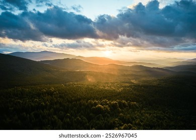 Aerial view breathtaking sunset over forest. Sun's rays break through dramatic clouds, casting golden glow across rolling hills and valleys. Distant mountains creating serene and picturesque scene. - Powered by Shutterstock