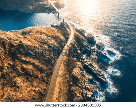 Aerial view of bridge, car, sea with waves and mountains at sunset in Lofoten Islands, Norway. Landscape with beautiful road, water, rocks and stones, golden sunlight. Top view from drone of highway