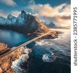 Aerial view of bridge, sea with waves and snowy mountains at sunset in Lofoten Islands, Norway. Landscape with beautiful road, water, rocks, blue sky with clouds and golden sunlight. Top drone view