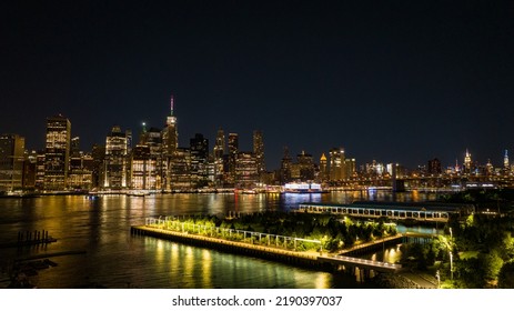 An Aerial View Of Brooklyn Bridge Park At Night With Lower Manhattan In The Background, Reflecting Lights Onto The Calm And Reflective East River.