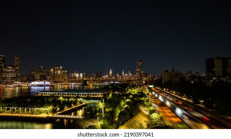 An Aerial View Of Brooklyn Bridge Park At Night With Lower Manhattan In The Background, Reflecting Lights Onto The Calm And Reflective East River.