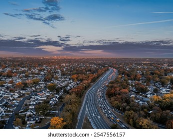 Aerial view of a busy highway with autumn foliage and suburban homes, leading to a distant city skyline at sunset. - Powered by Shutterstock