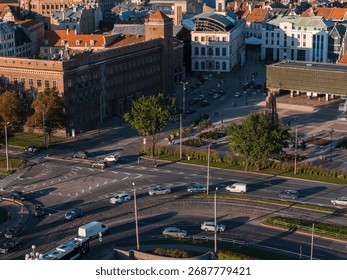 Aerial view of a busy intersection in Riga, Latvia, featuring a historic brick building with a clock tower, a modern glass roofed structure, and a park.