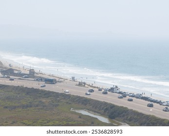 Aerial view of a busy road parallel to a San Diego beach, with parked cars, beachgoers, lifeguard towers, and a lush green area under a hazy sky. - Powered by Shutterstock