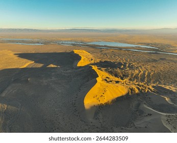 An aerial view captures the dramatic form of the Singing Barkhan sand dune casting a long shadow across the desert landscape at sunset, with distant water bodies