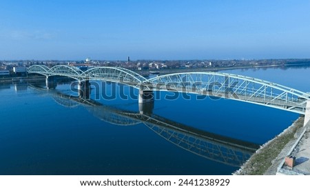 An aerial view captures the graceful arches of a steel bridge spanning across a tranquil river, its perfect reflection mirroring on the calm waters below.