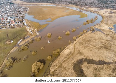 Aerial view captures a substantial spring river inundation near a small settlement, submerging fields and bushes with muddy water