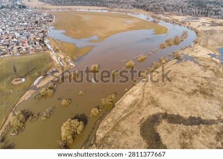 Aerial view captures a substantial spring river inundation near a small settlement, submerging fields and bushes with muddy water