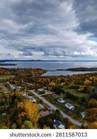 Aerial view capturing a tranquil waterside residential neighborhood. Autumn trees surround the homes as a dramatic, cloudy sky looms over the scene.