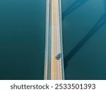 Aerial view of a car driving across a bridge, casting long shadows over calm blue water, creating a minimalist scene of travel and solitude.