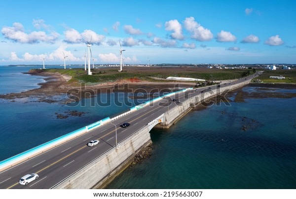 Aerial view of cars driving on a highway across\
the blue seawater and giant wind turbines standing by the coast in\
background on a sunny summer day, in Zhongtun, Baisha Township,\
Penghu County, Taiwan