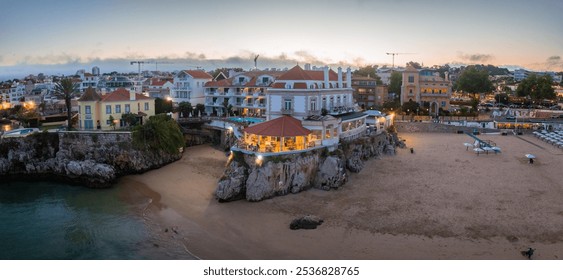 Aerial view of Cascais, Portugal, featuring a sandy beach, rocky cliffs, and a large red tiled building. The setting sun casts a warm glow over the scene. - Powered by Shutterstock