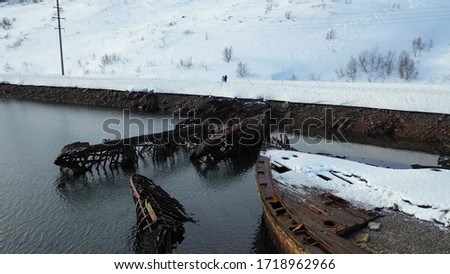 Aerial view of cemetery of old rusty ships near the snowy coast of cold sea in winter season. Footage. Flying over ruined boats and snow covered hills near calm sea.