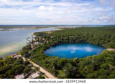 Aerial view of Cenote Azul surrounded by green jungle next to Bacalar lagoon with houses on a cloudy day
