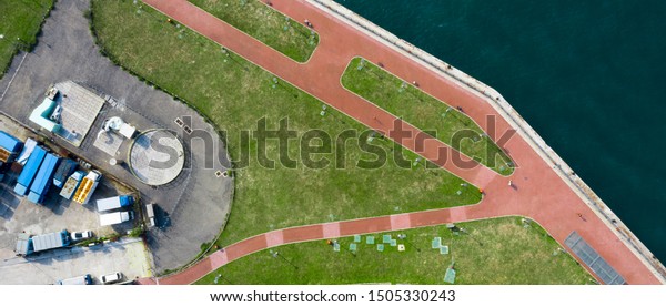 Aerial view of city park with running\
track near harbour front. Car park with blue trucks parking in the\
lot. Green grass and pink running track near the\
sea.