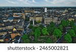 Aerial view of a cityscape featuring a mix of historic and modern buildings, a central monument, and lush green trees in a park area.