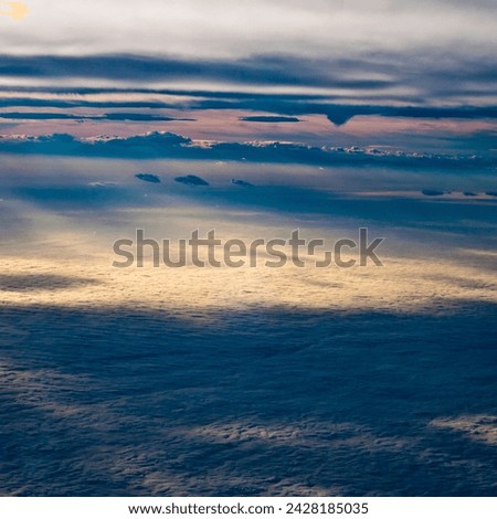 Aerial view of a cloudscape at dusk, showcasing layers of clouds with the setting sun casting a soft glow, illuminating the edges and creating shadows.