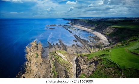 Aerial view of a coastal landscape with cliffs, rocky shoreline, and green fields under a partly cloudy sky.