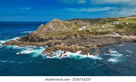 Aerial view of a coastal landscape featuring a rocky shore, rolling green hills, and a lighthouse on a hilltop. The ocean waves crash against the rocks, creating white foam. At Cape Cornwall, UK. - Powered by Shutterstock