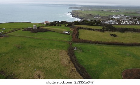 Aerial view of a coastal landscape with green fields, a cylindrical tower, and houses by the sea on a cloudy day. - Powered by Shutterstock