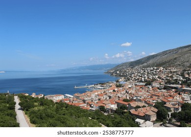 An aerial view of a coastal town with red-roofed buildings, nestled along a scenic coastline with blue waters and mountains in the background under a clear sky - Powered by Shutterstock