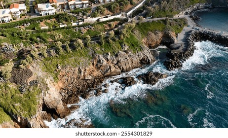 An aerial view of a coastal village perched above a rocky shoreline, featuring waves crashing against the cliffs and vibrant greenery along the rugged coast - Powered by Shutterstock