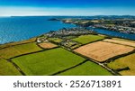 Aerial view of a coastal village surrounded by lush green fields and farmland. The blue sea stretches out in the background, with boats dotting the water. In Cornwall, UK.