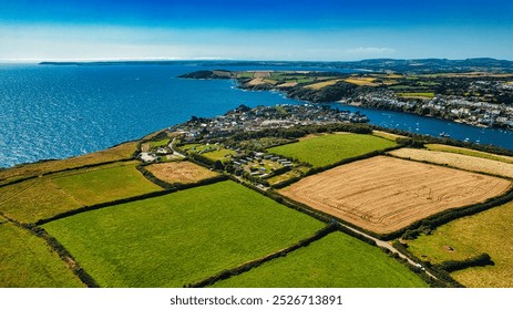 Aerial view of a coastal village surrounded by lush green fields and farmland. The blue sea stretches out in the background, with boats dotting the water. In Cornwall, UK. - Powered by Shutterstock