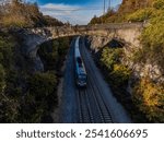 An aerial view of a commuter train approaching, taken with a drone on a sunny day in autumn. The train is in a deep ravine between a rock cliff and colorful trees.