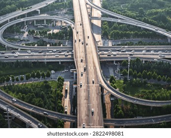 Aerial view of a complex highway interchange with sweeping circular ramps and a straight elevated main road. Strong pre-sunset sunlight reflects brightly off the asphalt, casting long shadows. Lush green landscapes and a ground-level transit stop surround this geometric urban infrastructure in Zhengzhou.