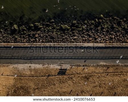 An aerial view of a concrete boardwalk off Lower Bay in Brooklyn, New York on a sunny day. The drone camera shows a quiet park with a paved walkway.