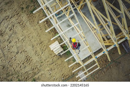 Aerial View Of Construction Worker In Yellow Hard Hat Working On Residential Building Wooden Roof Truss To Build A Frame For Roof Covering. Domestic Construction Industry Theme.