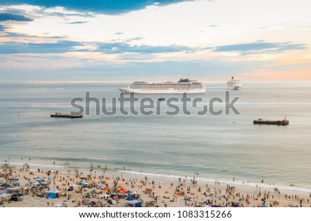 Aerial view of crowded beach with two large cruise ships on the horizon at sunset.