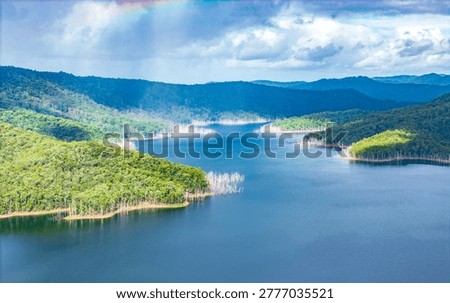 An aerial view of a deep blue mountain reservoir bordered by lush forests and stark, bleached dead trees under a cloudy sky.
