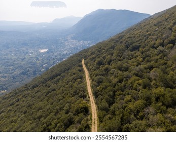 Aerial view of a dirt road winding through a lush green forest, leading down a mountainside towards a valley. - Powered by Shutterstock