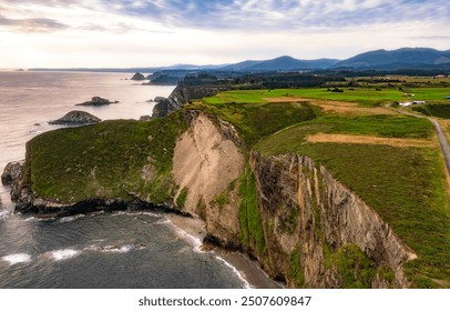 Aerial view of dramatic coastal cliffs with green fields, calm ocean waves, and distant mountains - Powered by Shutterstock