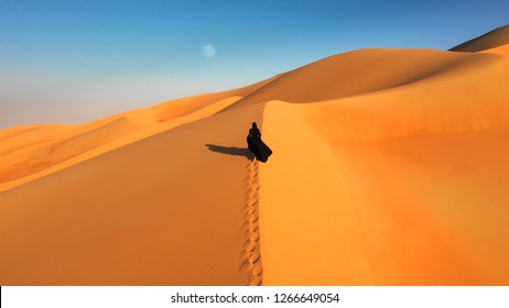 Aerial view from a drone flying next to a woman in abaya (United Arab Emirates traditional dress) walking on the dunes in the desert of the Empty Quarter. Abu Dhabi, UAE.
