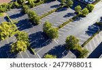 Aerial view of an empty parking lot, bordered by green trees and bushes, with clearly marked parking spaces and drive lanes.