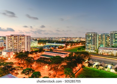 Aerial View Of Eunos HDB Next To MRT Station In Singapore At Twilight. Colorful City Lights, Electric Train, Car Headlights In Traffic. Neighborhood Faculties Car Park And Green Garden At The Center.