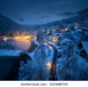 Aerial view of fairy town in snow, road, forest, Jasna lake and houses with lights at night in winter. Top view from drone of mountain village, pond, illumination, snowy pine trees at dusk in Slovenia - Powered by Shutterstock