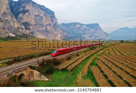 Aerial view of a fast train dashing through the beautiful landscape of vineyard fields on a sunny autumn day and a rugged mountain wall dominating the background, in Mezzocorona, Trentino, Italy