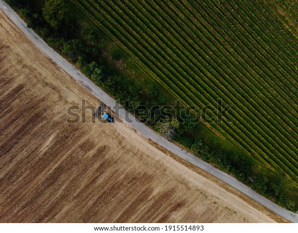 Aerial view of field in which tractor plows.Blue\
tillage equipment and blue tractor are clearly visible on  brown\
background of earth.Road divides scape in two halves with green\
color in other one.