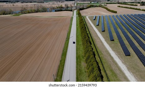 An Aerial View Following A Blue Car Driving On A Country Road Between A Solar Farm And Field, Seen On A Sunny Day.